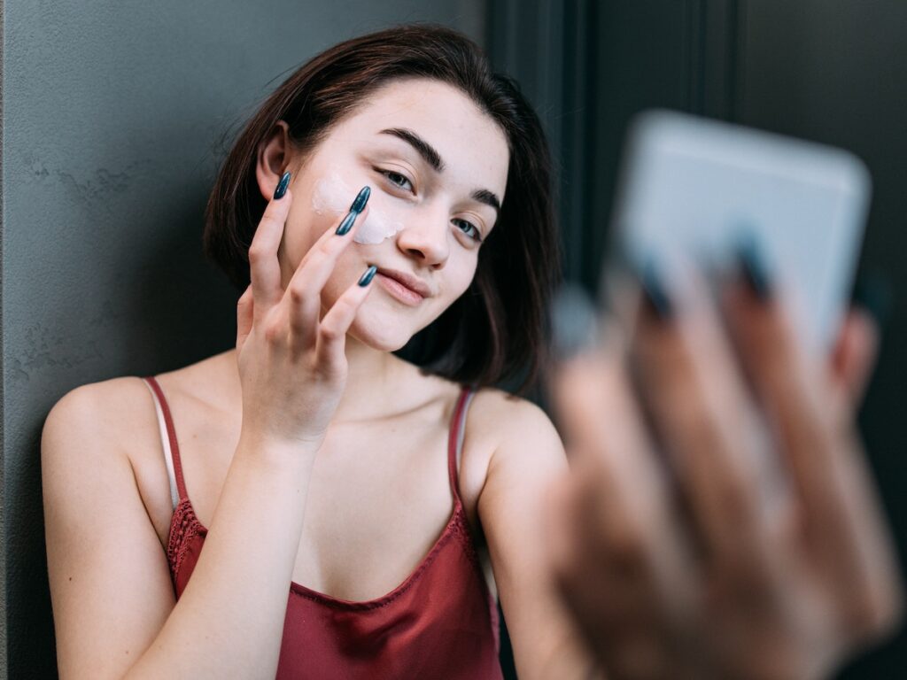 Radiant young woman in a maroon top applies Salicylic Acid cream, promoting clear, healthy skin against a dark background.